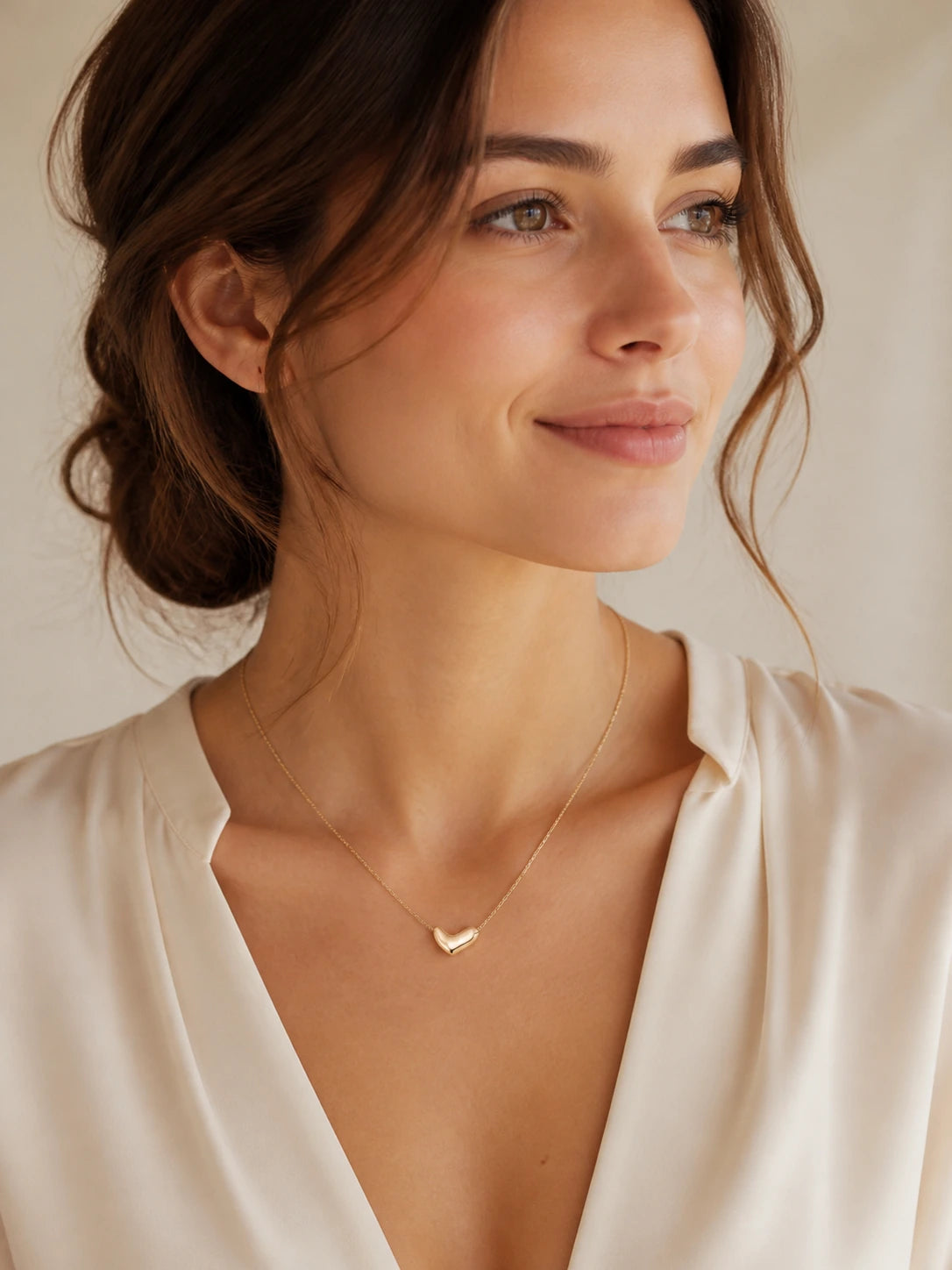 Woman in an ivory blouse wearing the gold Amour Heart Necklace in a softly lit studio setting.