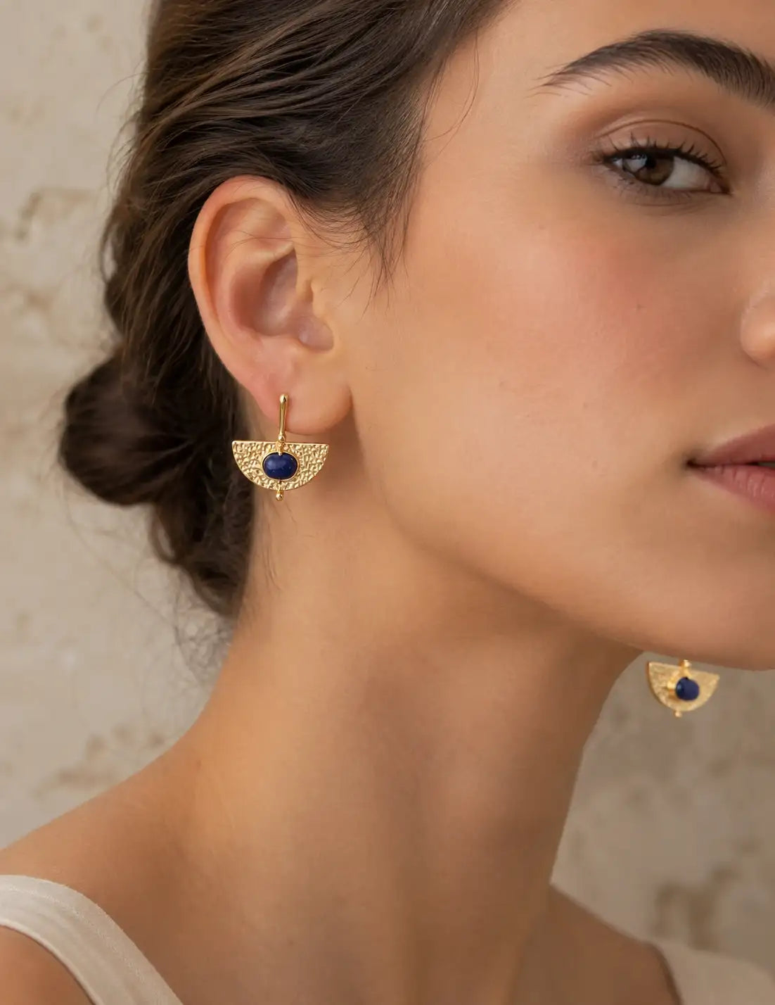 Close-up of a woman wearing gold dangle earrings with a central dark blue oval stone, shown against a soft beige textured background.