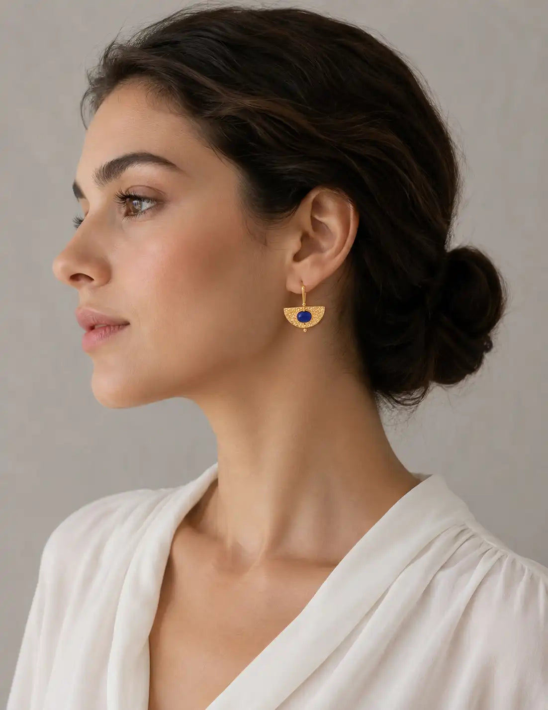 Woman wearing gold earrings with a deep blue stone, styled with a white blouse in a minimalist jewellery portrait.