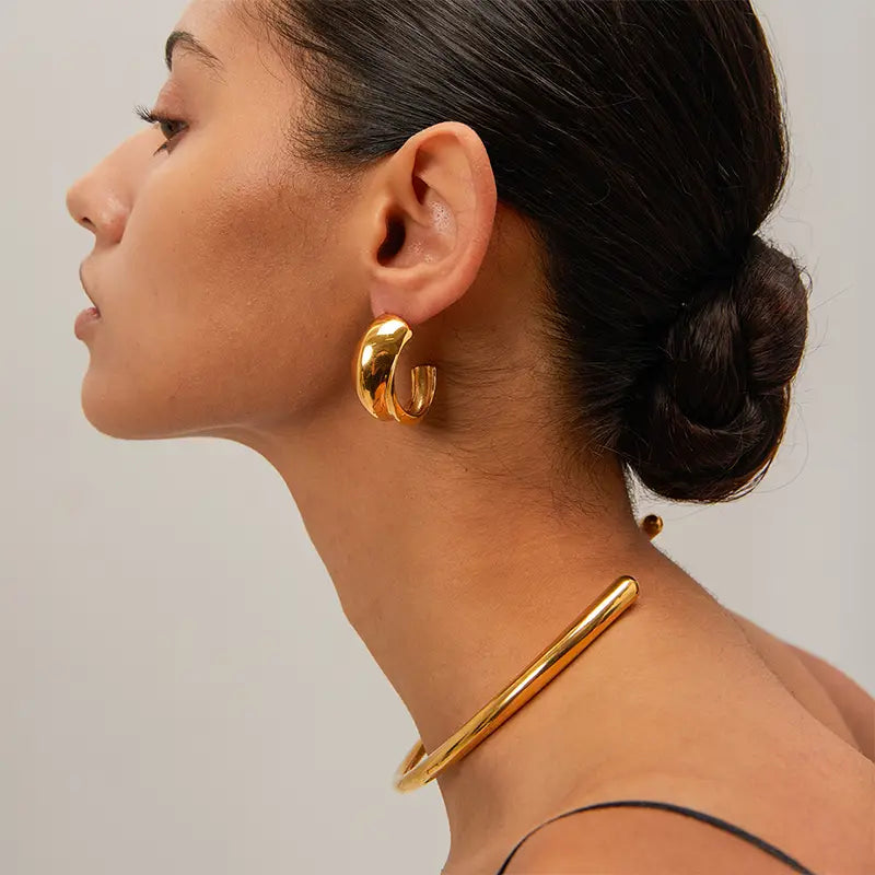 A close-up image of a person wearing gold-colored hoop earrings with a sleek C-shaped design, showing the earrings' gold plating and the hoop's width and elegance.
