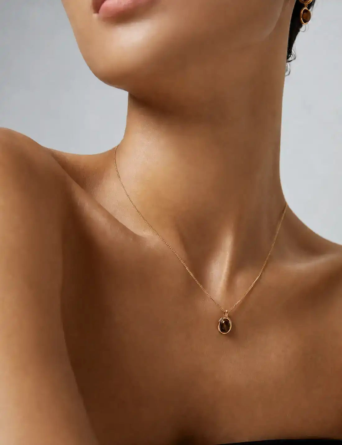 Model wearing a delicate gold necklace with an oval tiger’s eye pendant and matching earrings