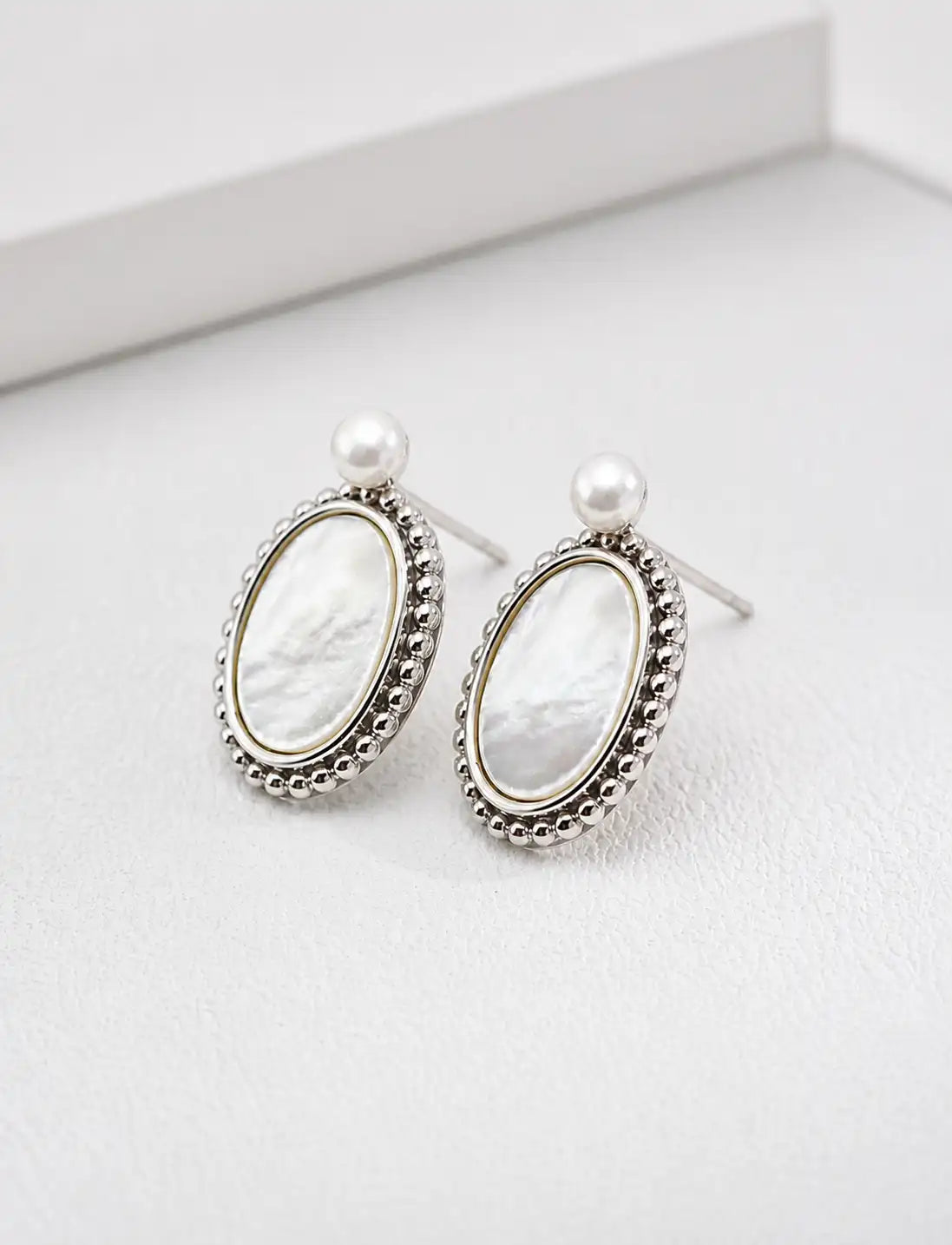A pair of silver-toned oval drop earrings featuring a white mother-of-pearl center, a small white pearl stud above, and a beaded border, displayed on a clean white surface.