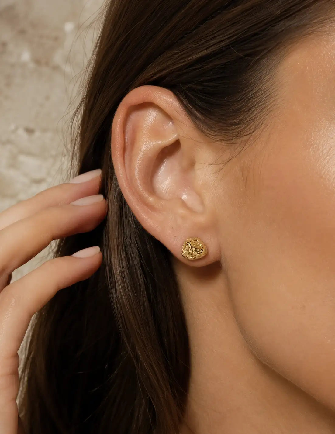 Side profile of a woman wearing textured gold stud earrings, photographed in warm natural light with a minimalist cream background.