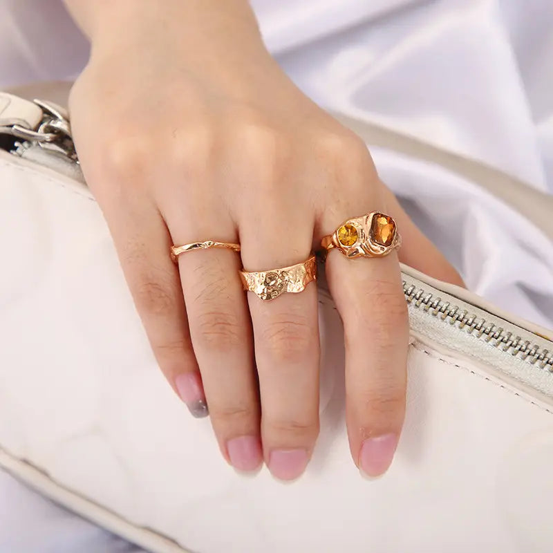 Trio gold rings with amber stone, hammered and wavy designs on hand holding white clutch.