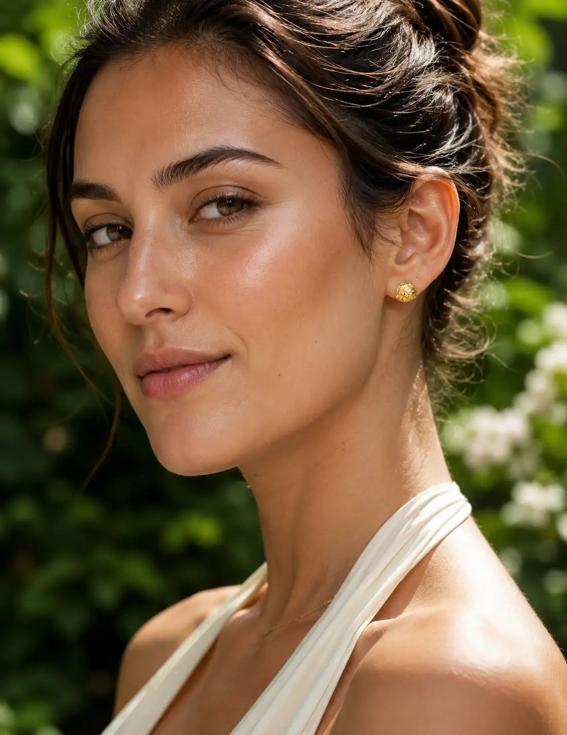 Close-up portrait of a woman wearing small textured gold stud earrings, styled with a cream outfit and natural garden background for an elegant everyday jewellery look.