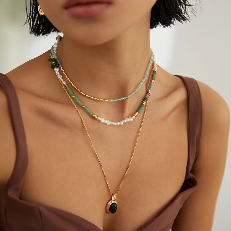 Woman wearing green crystal necklace and long gold necklace with black pendant over brown top.